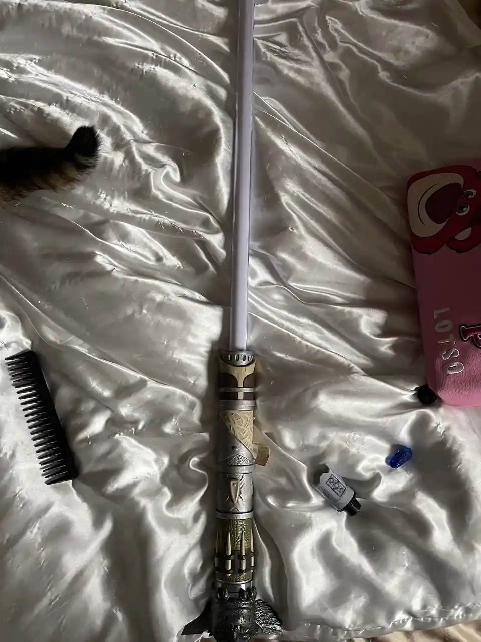 A white lightsaber with a detailed hilt lies on a rumpled white satin sheet, surrounded by everyday items including a black…