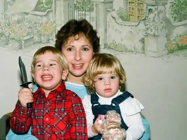 A vintage family photo shows a smiling woman with two young children; the boy on the left holds a large toy knife and grins…