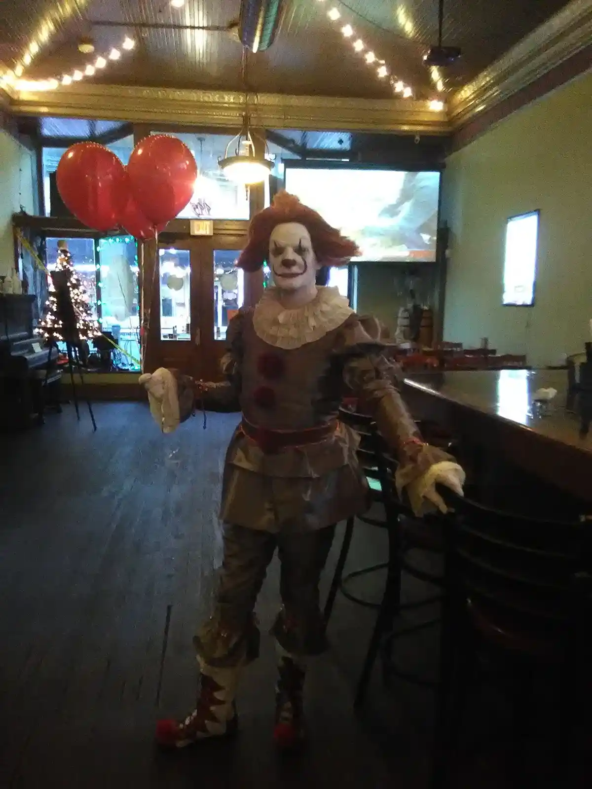 A person dressed as Pennywise the clown from IT stands inside a dimly lit bar, leaning on a barstool. Behind them, red…