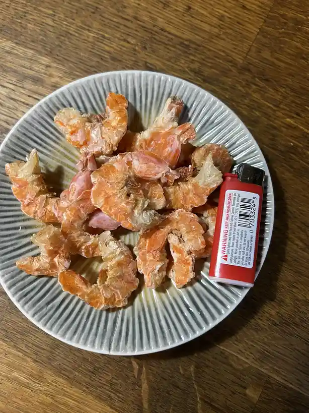A white plate with ridged edges holds dried, shriveled shrimp on a wooden table, next to a red lighter with a warning label…