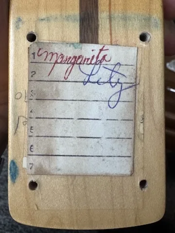 A close-up of a wooden guitar body with a handwritten note taped to it. The note has lines numbered 1 through 7, with…