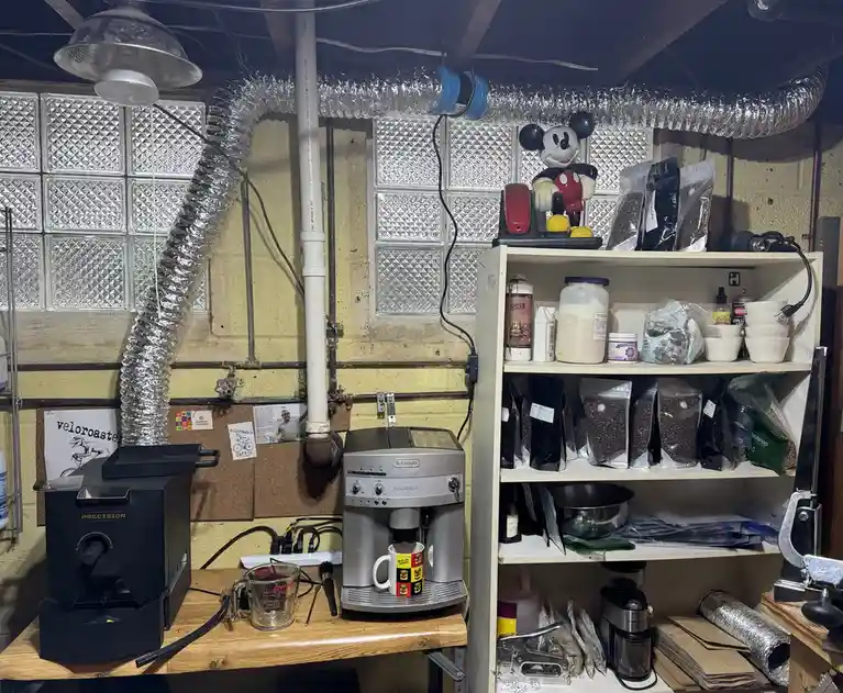 A cluttered basement coffee station features a DeLonghi Magnifica espresso machine brewing into a mug, next to a black…