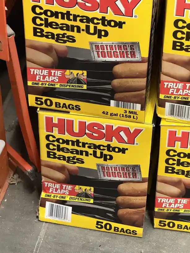 A close-up shot of yellow Husky brand Contractor Clean-Up Bags boxes stacked on a store floor, featuring bold red and black…