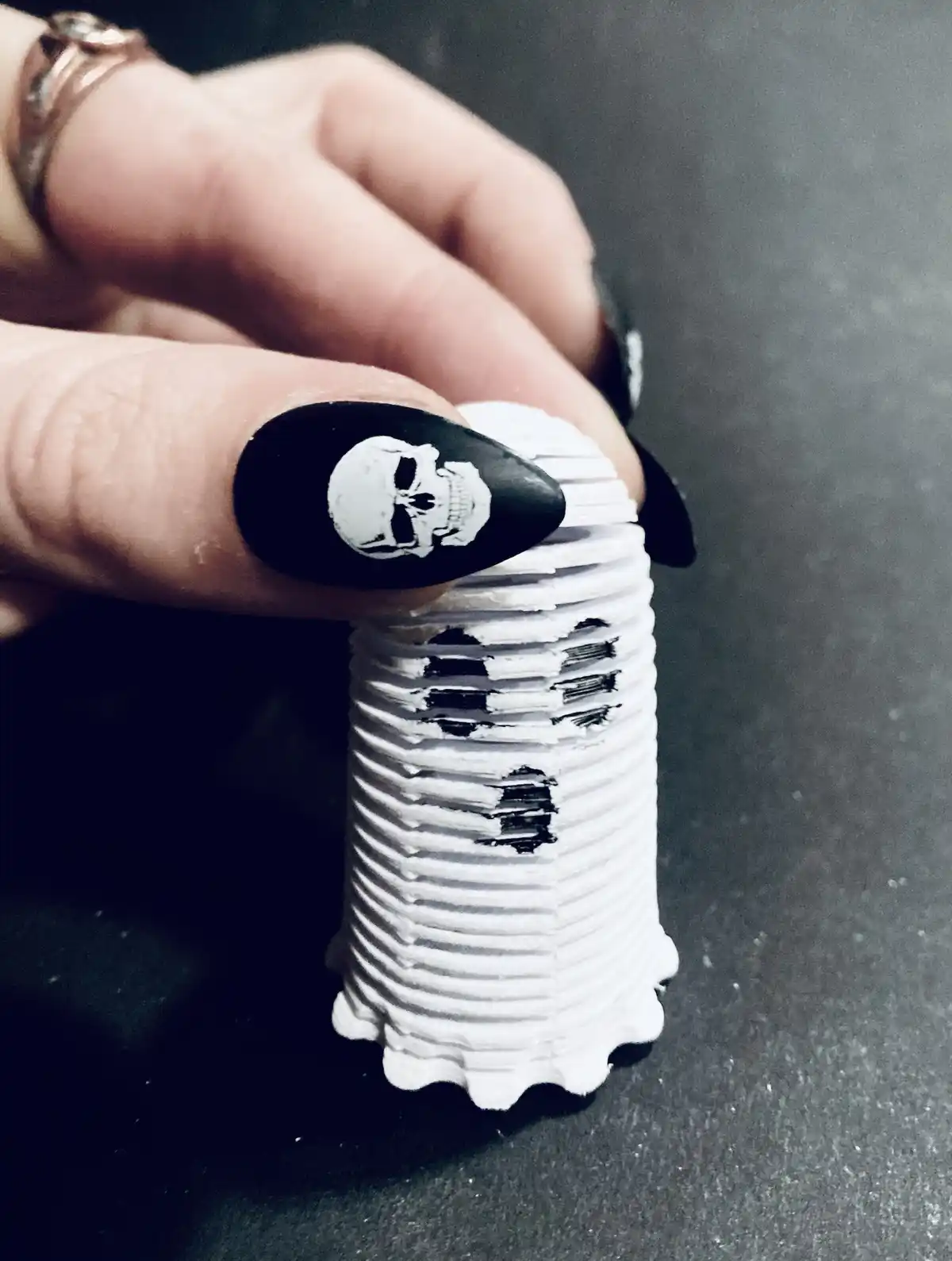 A hand with black coffin-shaped nails, one decorated with a white skull, holds a tall stack of white poker chips that have…