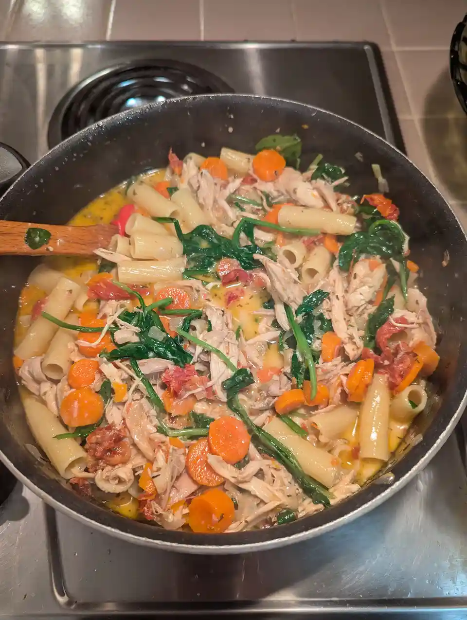 A close-up photo shows a black skillet on a stovetop filled with cooked pasta, shredded chicken, chopped carrots, wilted…