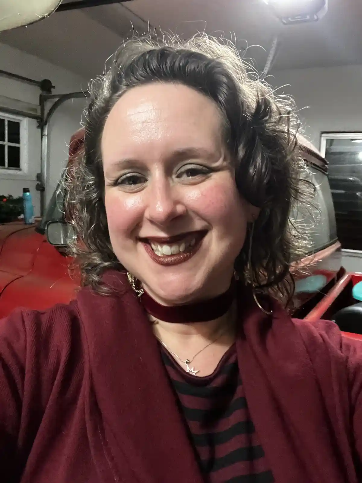 A woman with curly hair and a wide smile takes a selfie in a garage, wearing a red cardigan over a striped shirt and a red…