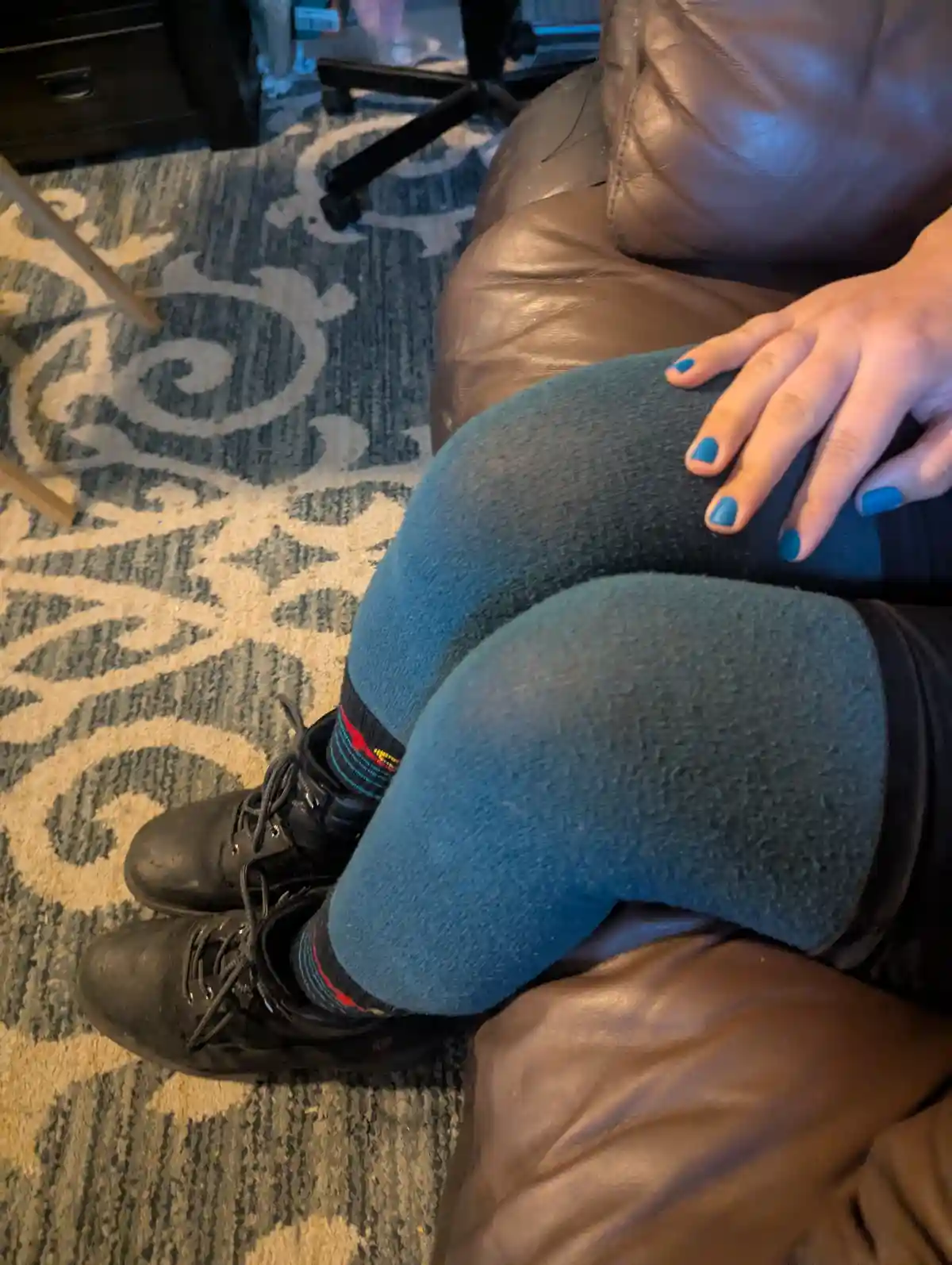 A person is sitting on a brown leather couch with their legs crossed, wearing blue fuzzy leggings, black boots, and colorful…