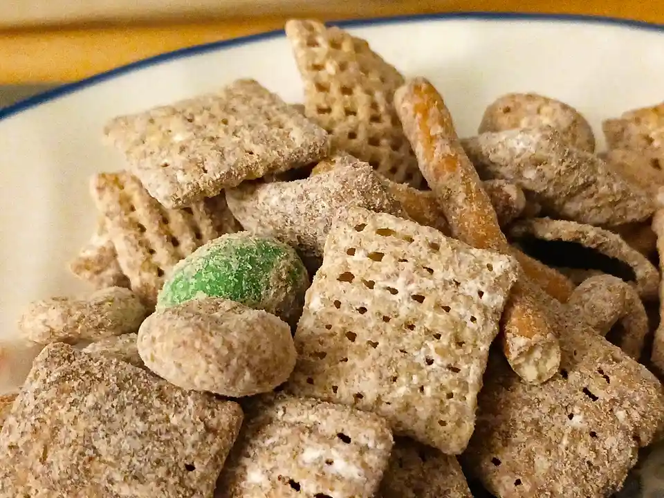 A close-up photo shows a white bowl with a blue rim filled with a snack mix containing square cereal pieces, pretzel sticks…