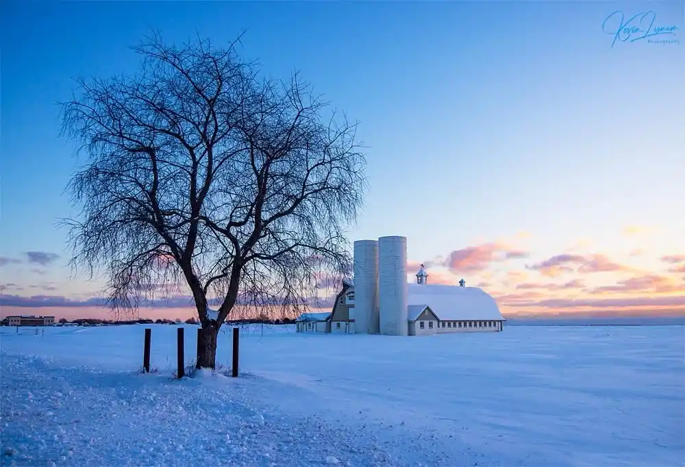 A serene winter landscape features a snow-covered farm with silos and a barn under a twilight sky with pink and blue hues. A…