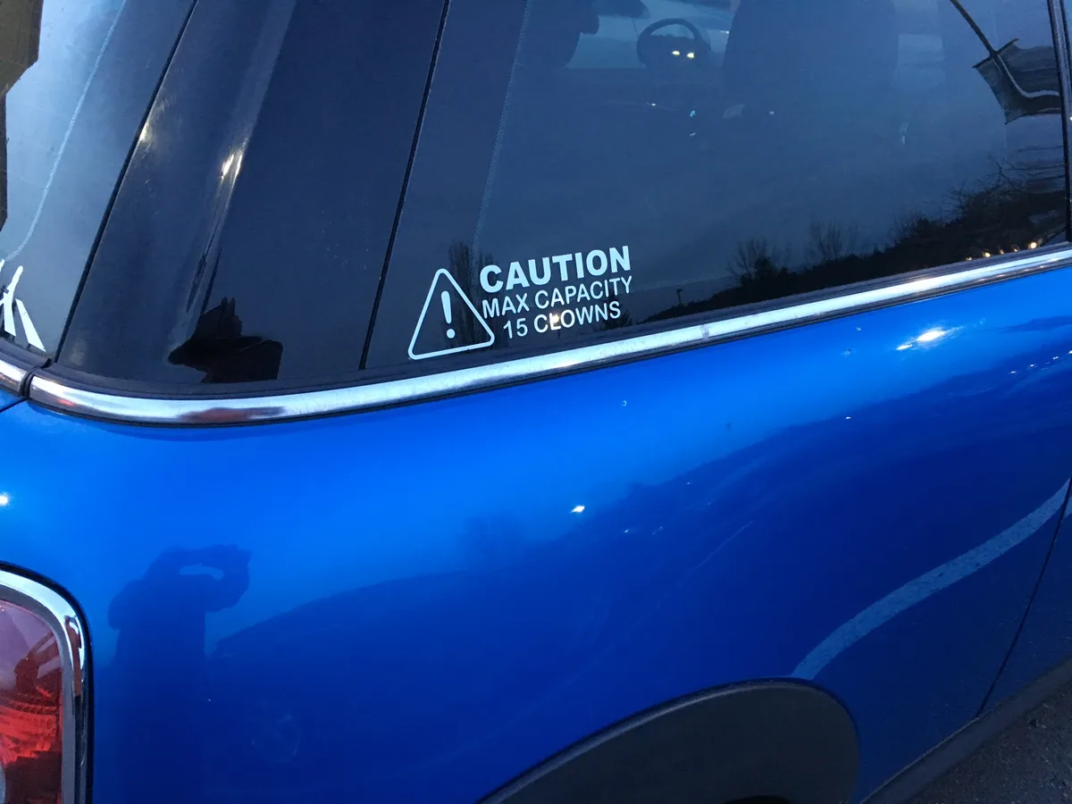 A close-up of a blue car's rear passenger window shows a white warning sticker with a triangular caution symbol and text…