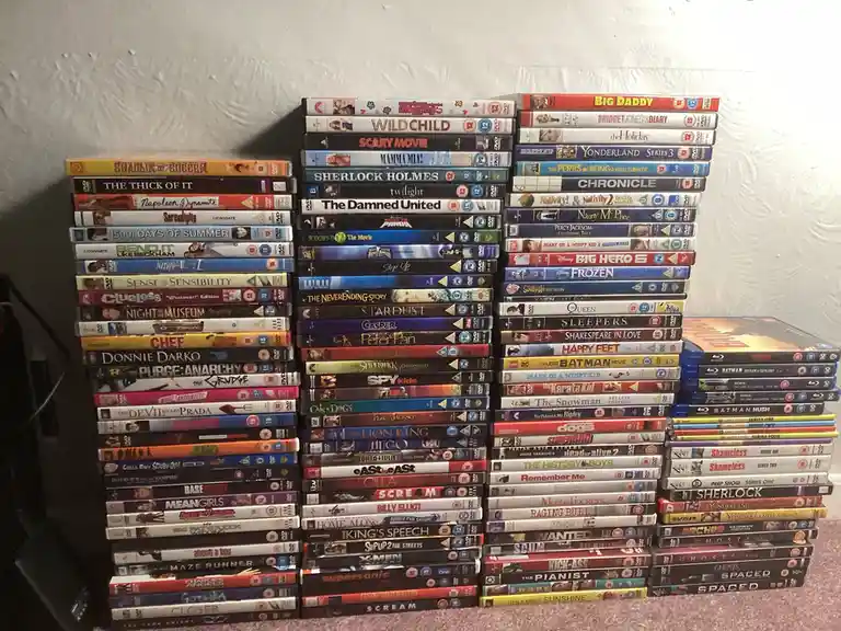 The image shows multiple tall stacks of DVD cases arranged on a carpeted floor against a white wall, featuring titles like…