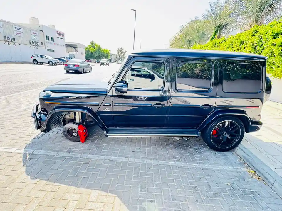 A black Mercedes-Benz G-Wagen is parked on a paved area with its front left wheel removed, exposing the red brake caliper…