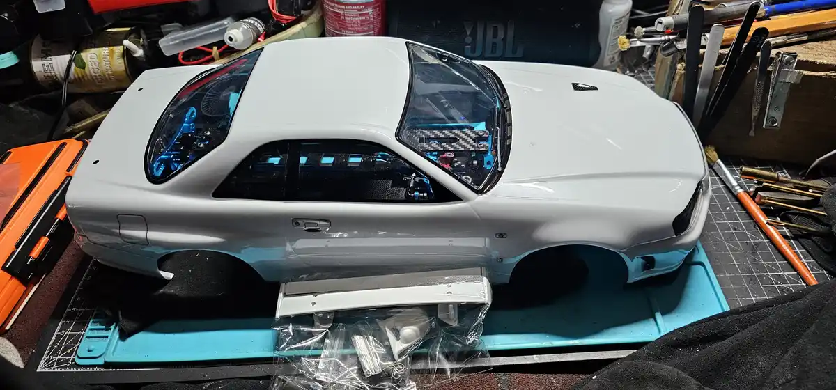 A silver 1:10 scale radio-controlled model of a Nissan Skyline R32 is shown from a side angle, its bodywork freshly painted…