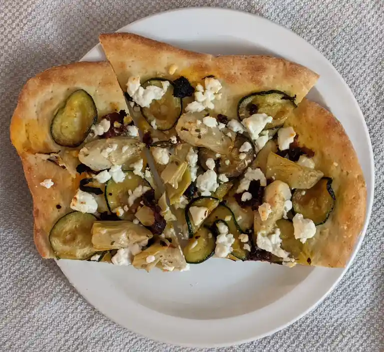 A close-up photo shows two slices of a rectangular flatbread pizza on a white plate, topped with roasted zucchini, artichoke…