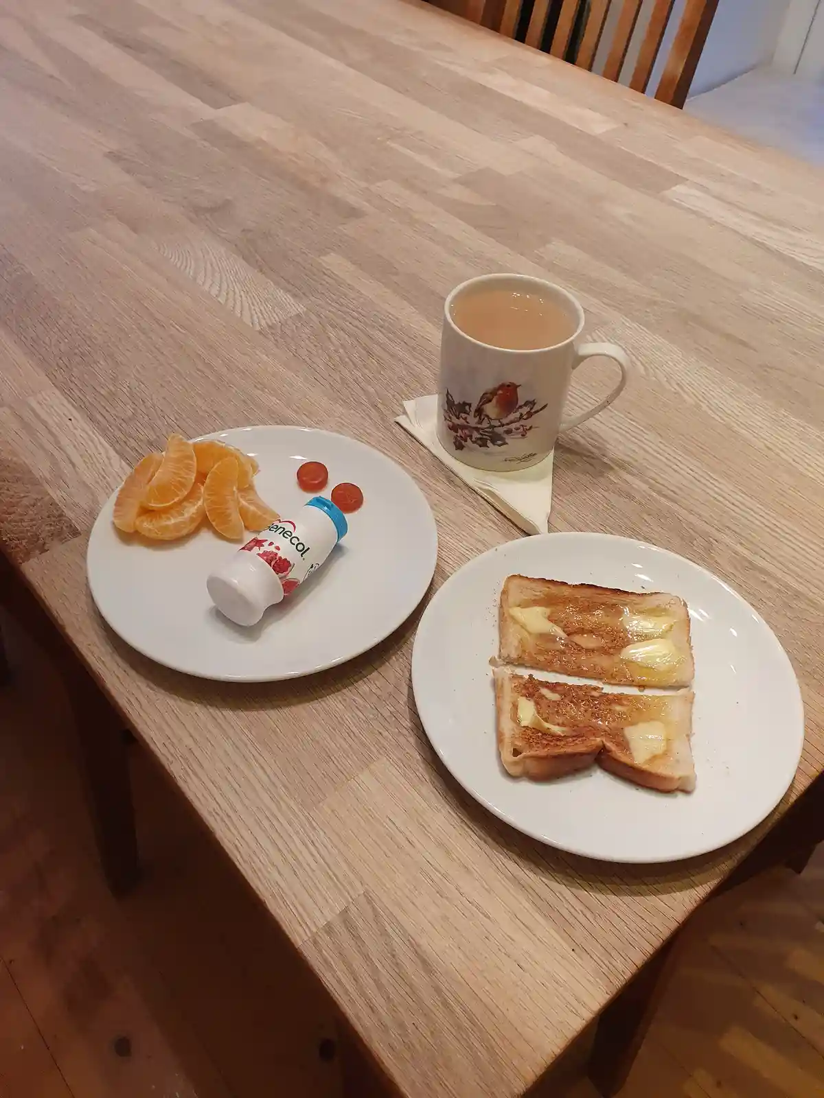 A wooden table holds a breakfast spread: one plate with buttered toast, another with orange segments, two cherry tomatoes…