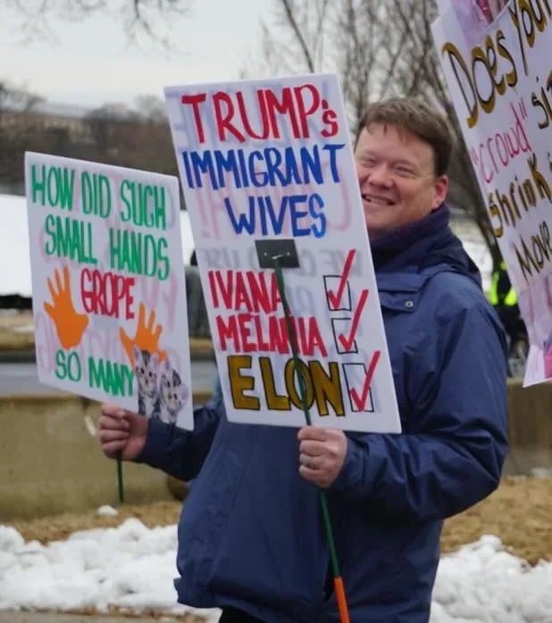 A man in a blue jacket smiles while holding two protest signs at an outdoor rally with snow on the ground. One sign reads…