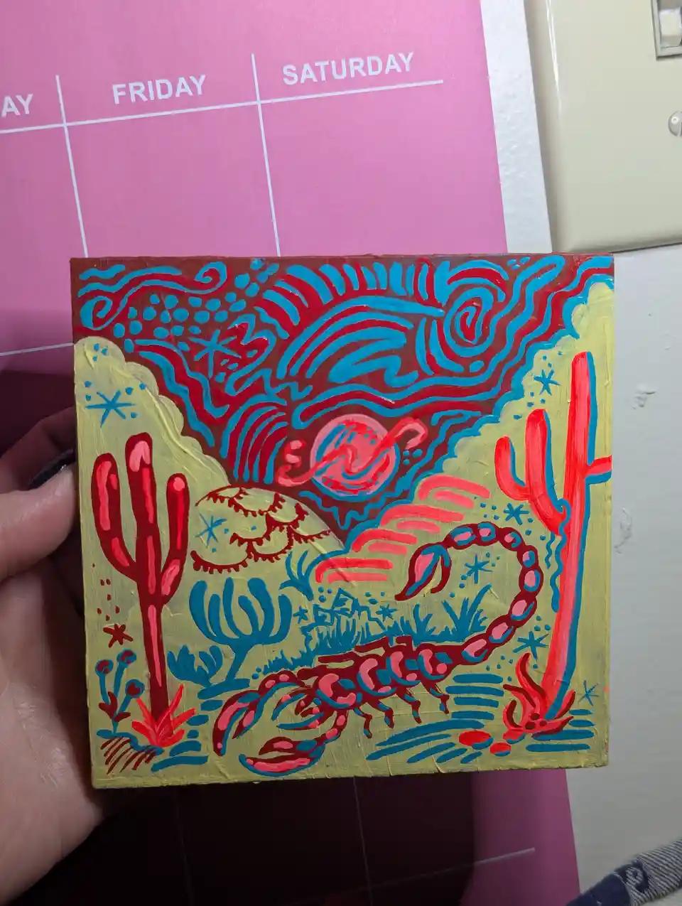 A hand holds a small, colorful painting depicting a desert scene with red cacti, a large red scorpion, and a stylized sun or…