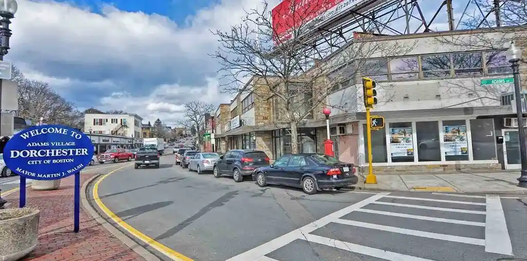 A street scene in Adams Village, Dorchester, Boston, featuring a blue welcome sign for the neighborhood and city, parked…