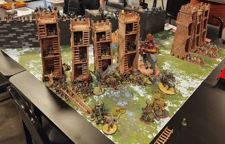 A tabletop wargaming scene shows numerous miniature figures and wooden siege towers attacking a fortified wall. The overlaid…