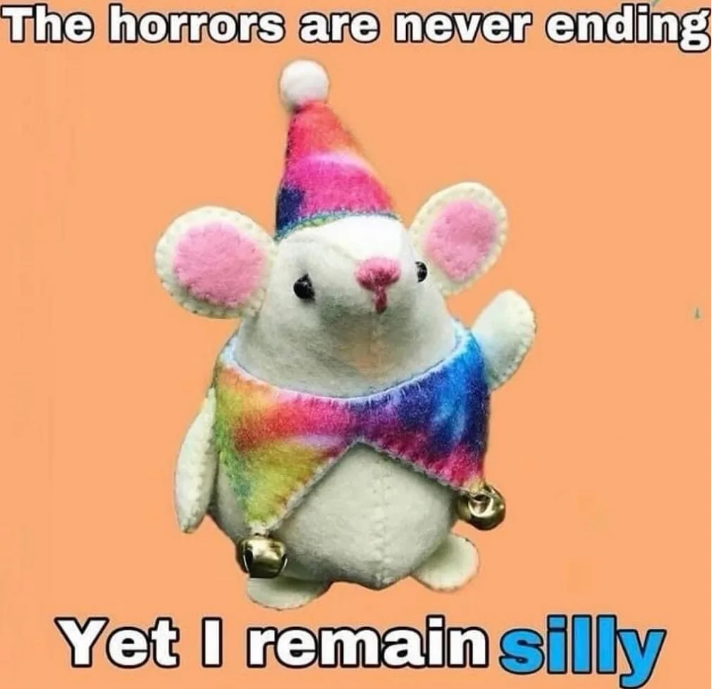 A plush white mouse toy wearing a rainbow tie-dye party hat and matching scarf with jingle bells, set against a solid peach…