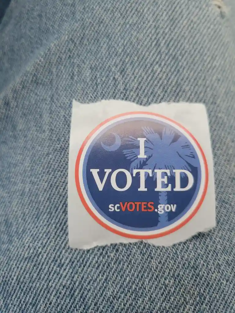 A circular 'I VOTED' sticker from scVOTES.gov is stuck on a pair of blue denim jeans. The sticker has a blue background with…