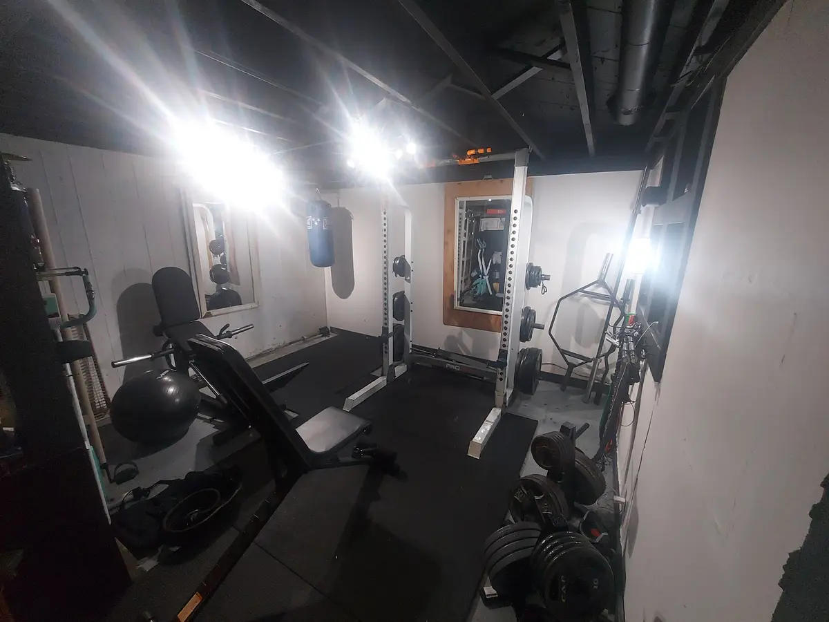 A dimly lit basement gym filled with weightlifting equipment including a squat rack, dumbbells, a bench, and a punching bag…