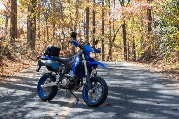 A blue adventure-style motorcycle is parked on a paved road in a forest with autumn foliage. The bike has a black seat, blue…