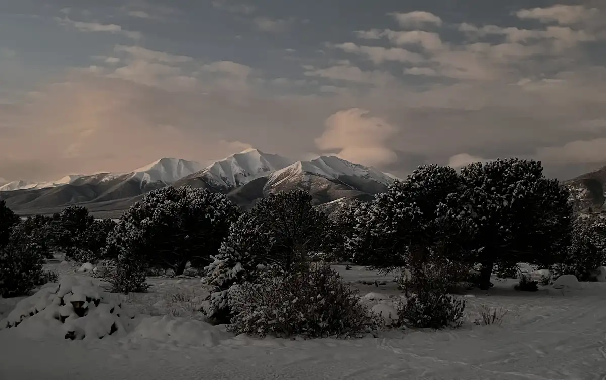 A serene winter landscape shows snow-covered evergreen trees in the foreground with majestic snow-capped mountains in the…