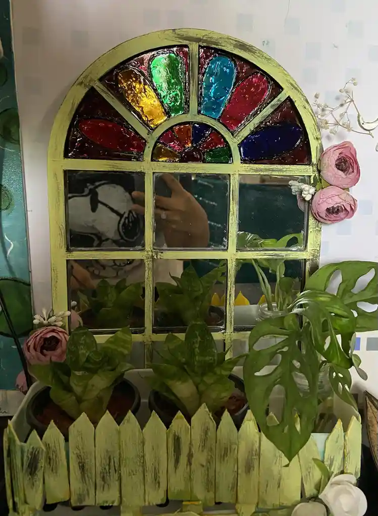 A decorative, arched stained-glass window mirror with a yellow frame reflects a person holding a phone, surrounded by potted…