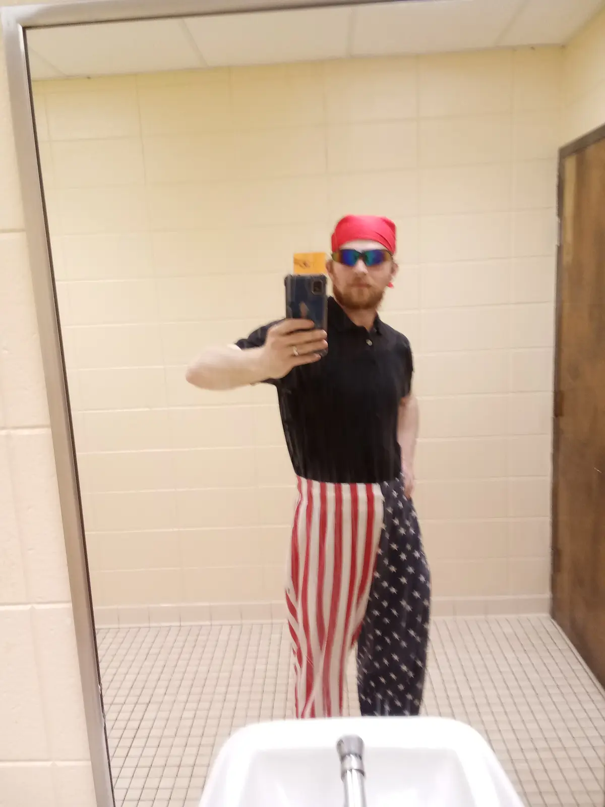 A man with a red bandana and sunglasses takes a mirror selfie in a public restroom, wearing pants split down the middle to…