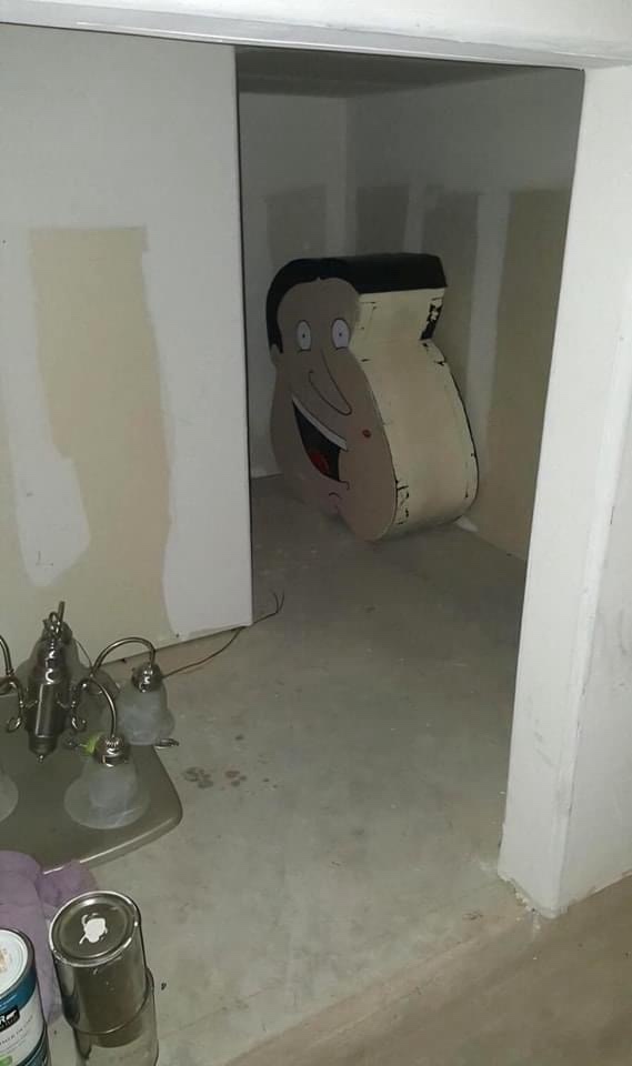 A large, cartoonish cutout of a smiling man with a black hairline and wide eyes leans against a wall in an unfinished room…