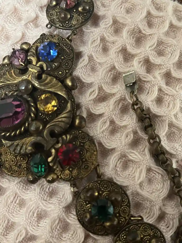 A close-up photo shows an ornate, vintage-style necklace with multiple colorful rhinestones set in brass-colored metalwork…