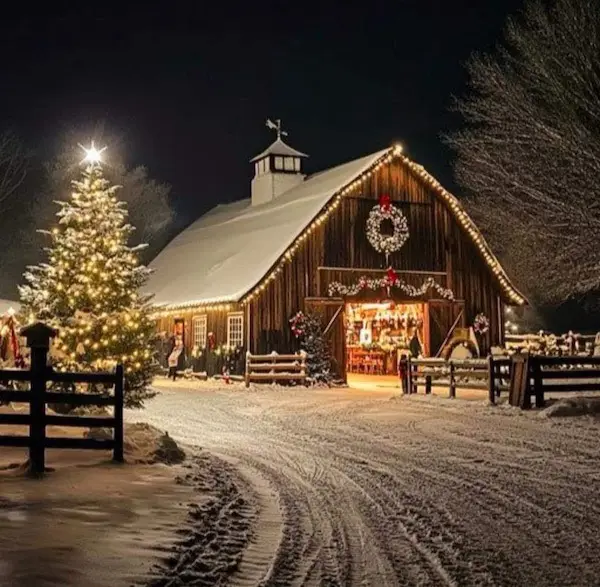 a snowy nighttime scene of a rustic wooden barn decorated with string lights, a wreath, and garlands, with a large lit…