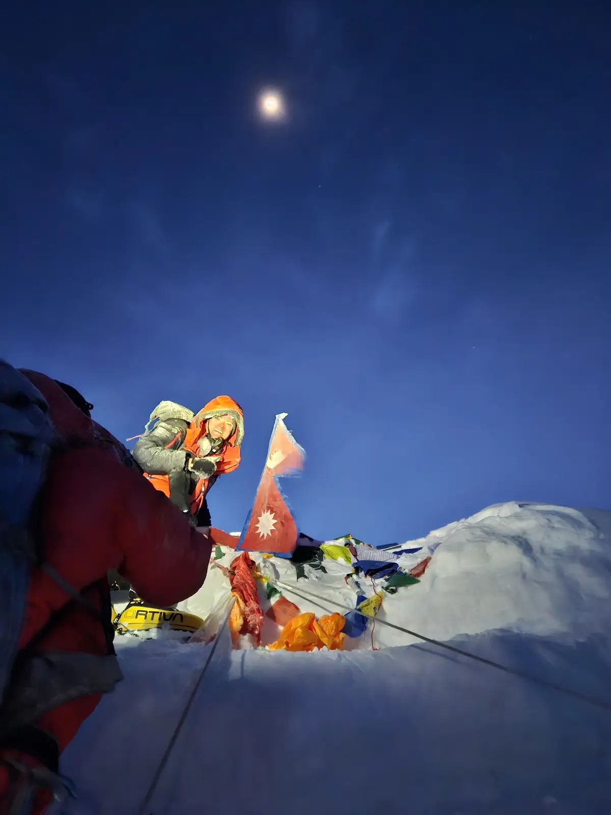 A climber in an orange parka and helmet smiles triumphantly atop a snowy peak under a bright moon, with a Nepalese flag and…