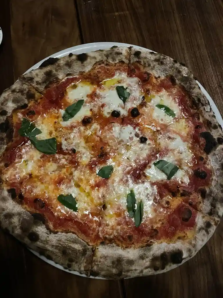 A close-up photo of a freshly baked Neapolitan-style pizza on a white plate, resting on a wooden table. The pizza has a…