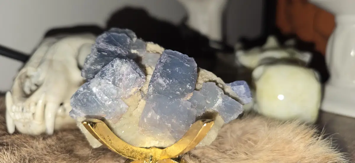 A close-up photo shows a blue and white mineral crystal cluster mounted on a golden crescent-shaped stand, resting on a…
