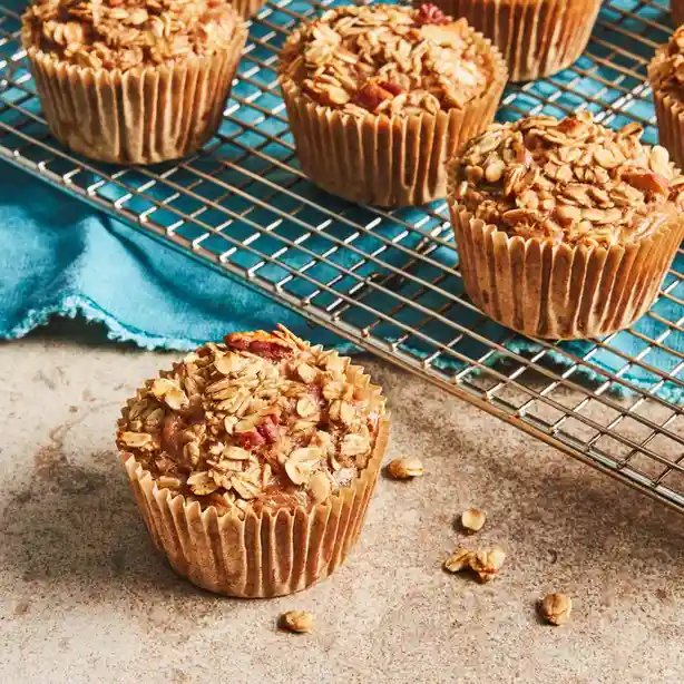 Several golden-brown muffins topped with oats and what appear to be dried cranberries sit on a wire cooling rack and a…