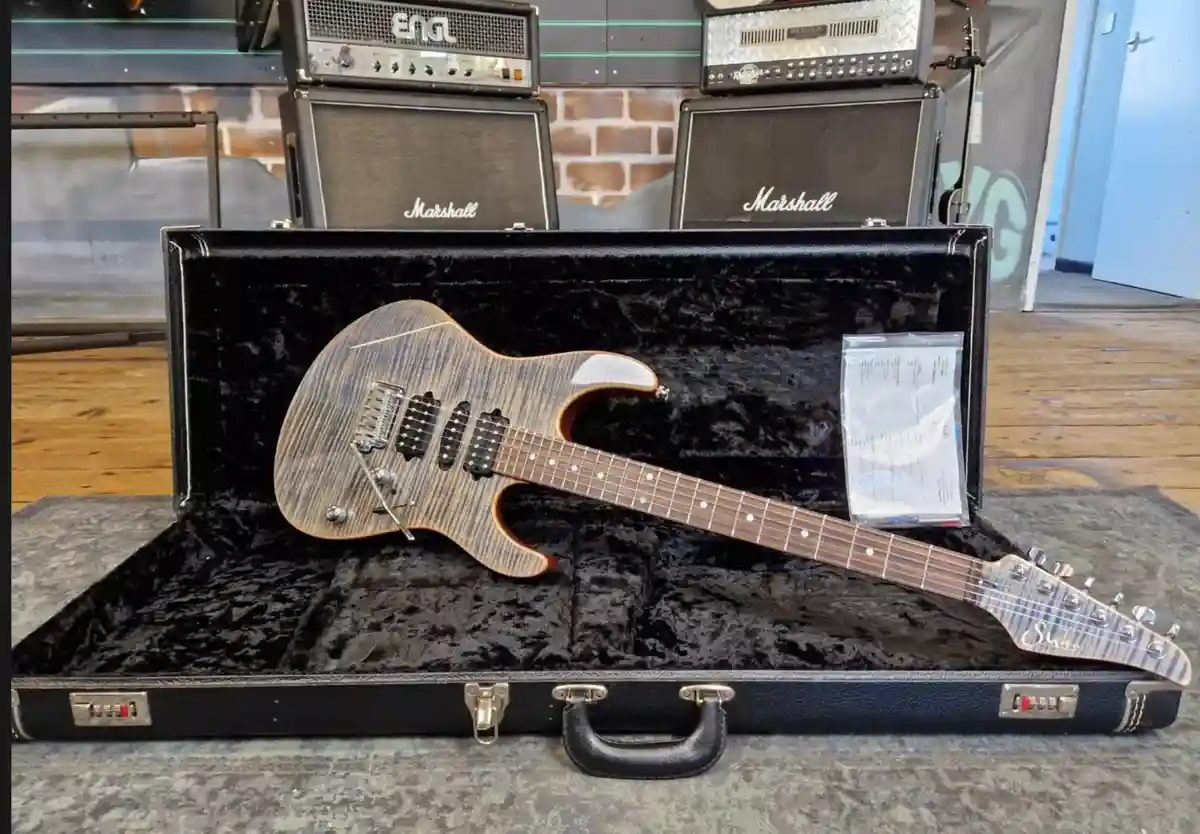 A Suhr electric guitar with a distinctive wood grain finish rests inside an open black hardshell case lined with black…