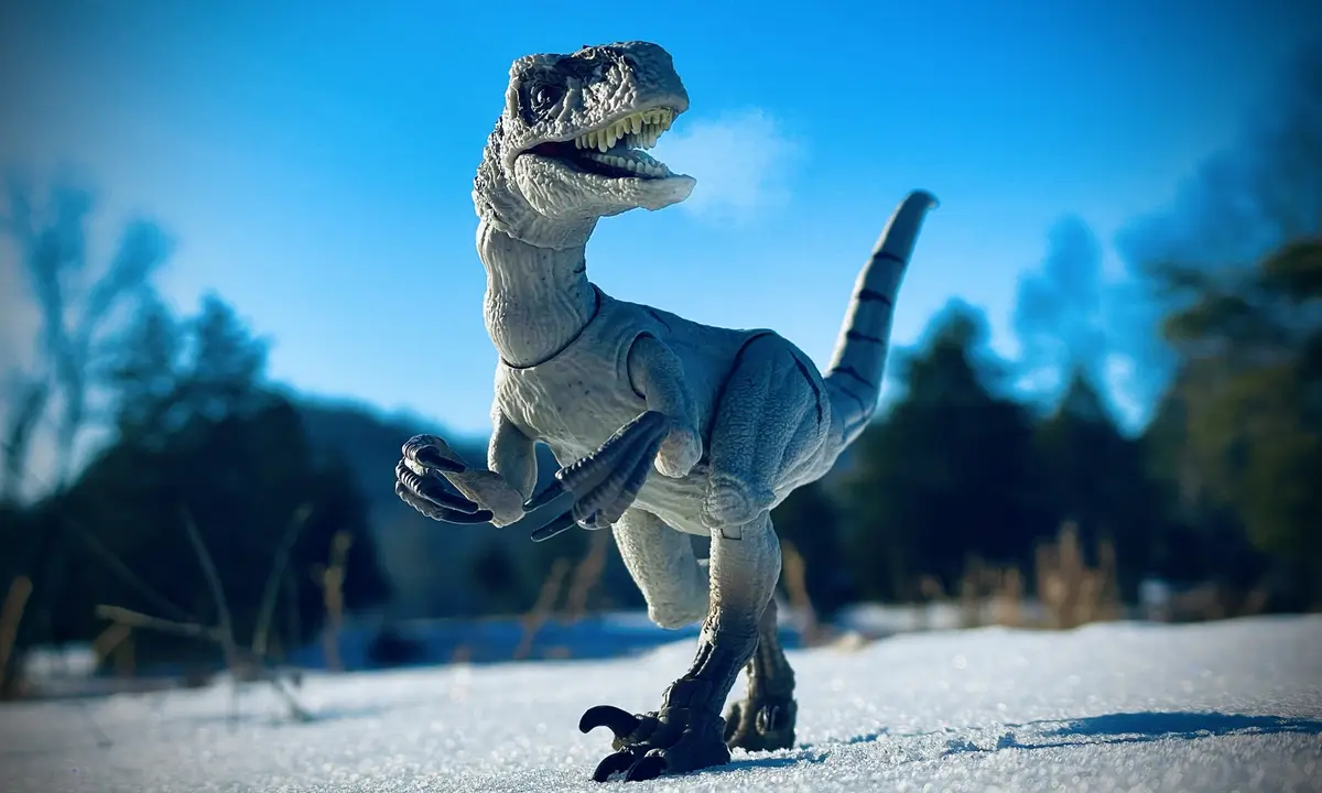 A plastic toy raptor stands in a snowy outdoor setting under a clear blue sky, its mouth open and claws visible. The…