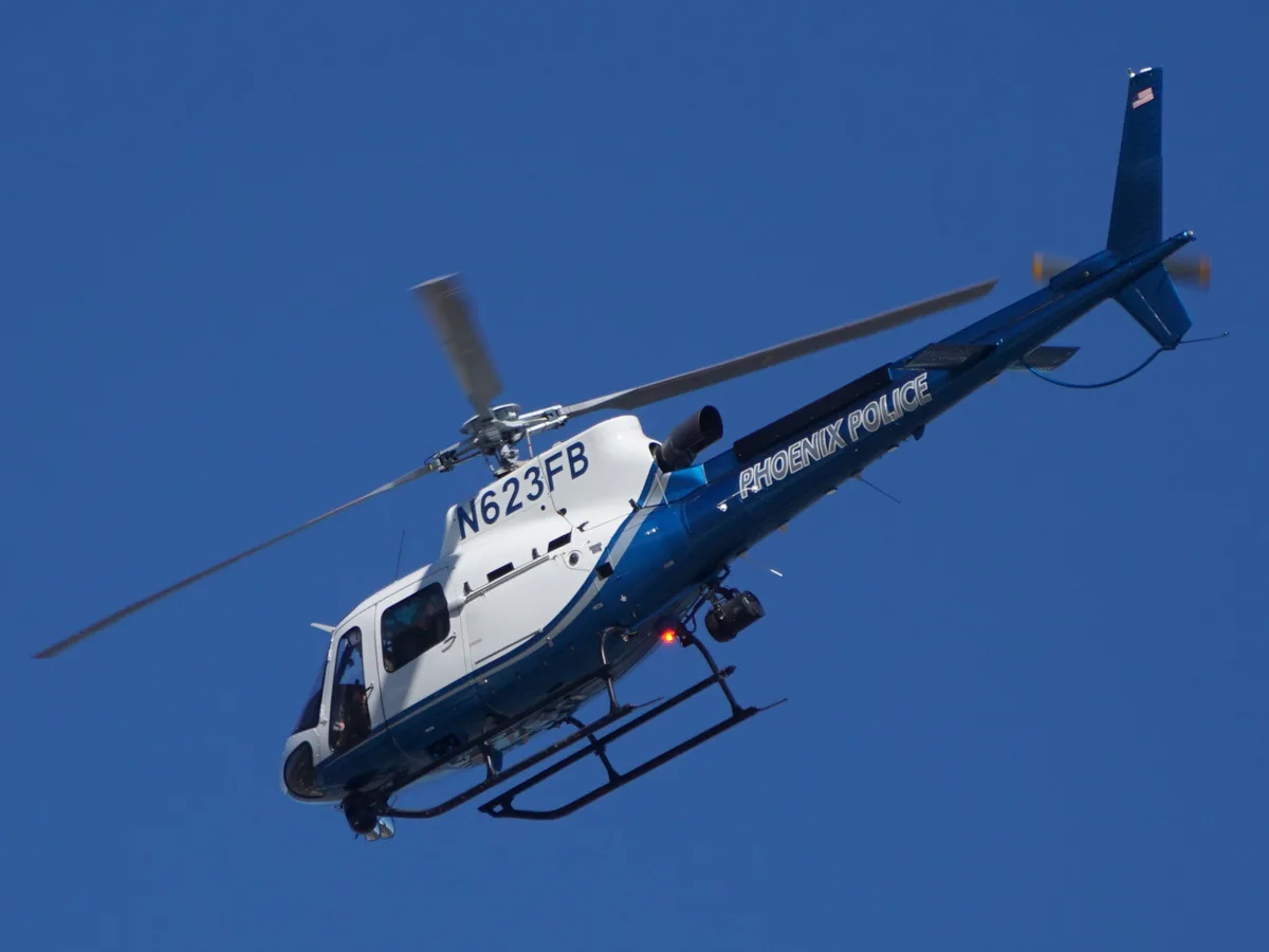 A blue and white Phoenix Police helicopter with registration N623FB is flying against a clear blue sky. The text 'phoenix…