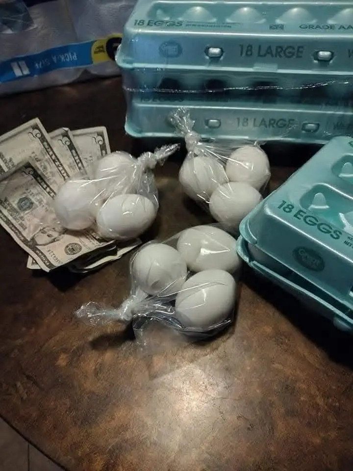 A wooden table holds several clear plastic bags filled with white eggs, next to stacks of US dollar bills and blue egg…