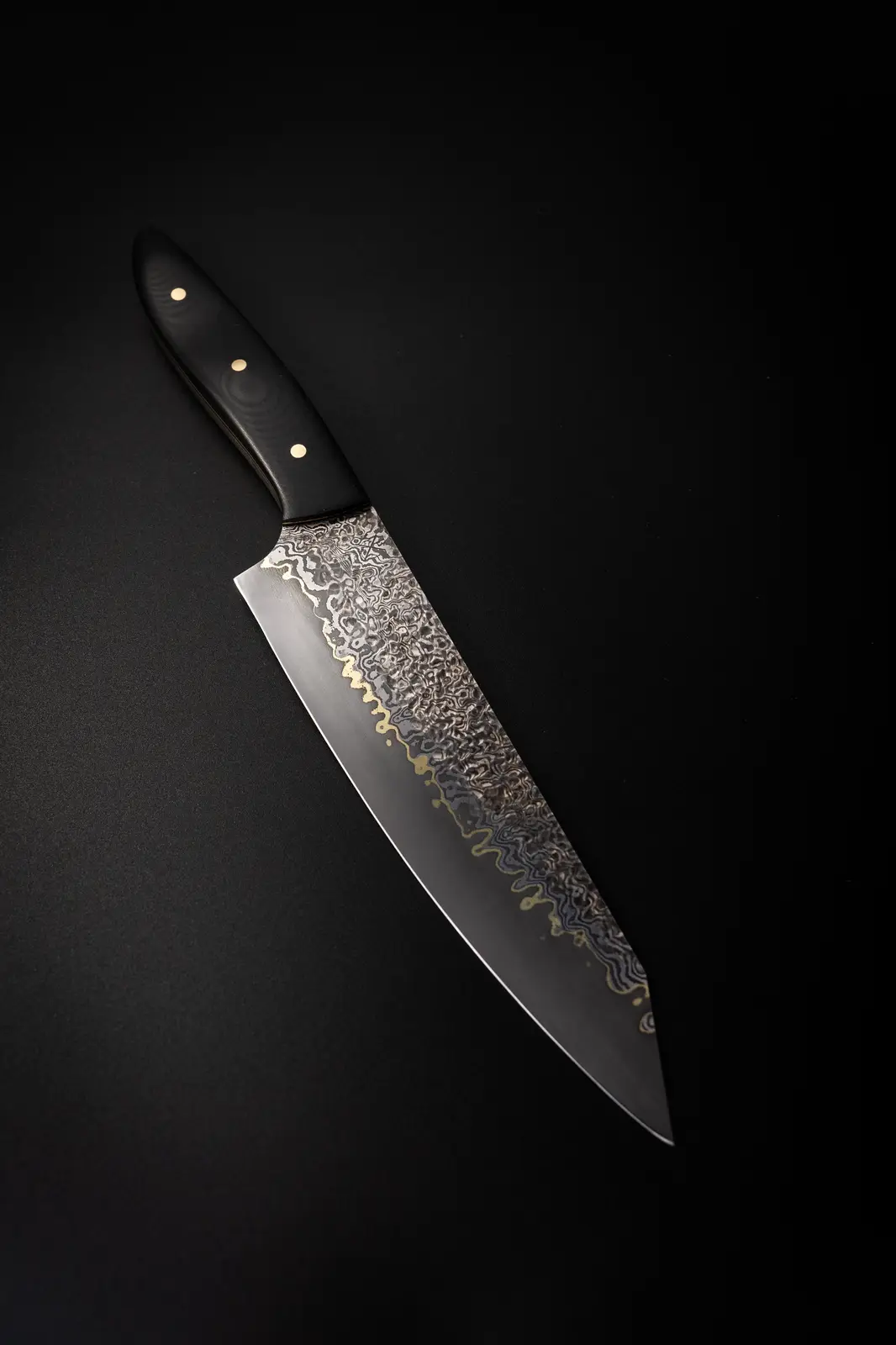 a high-quality chef's knife with a black handle and three brass rivets lies diagonally on a black surface. The blade…