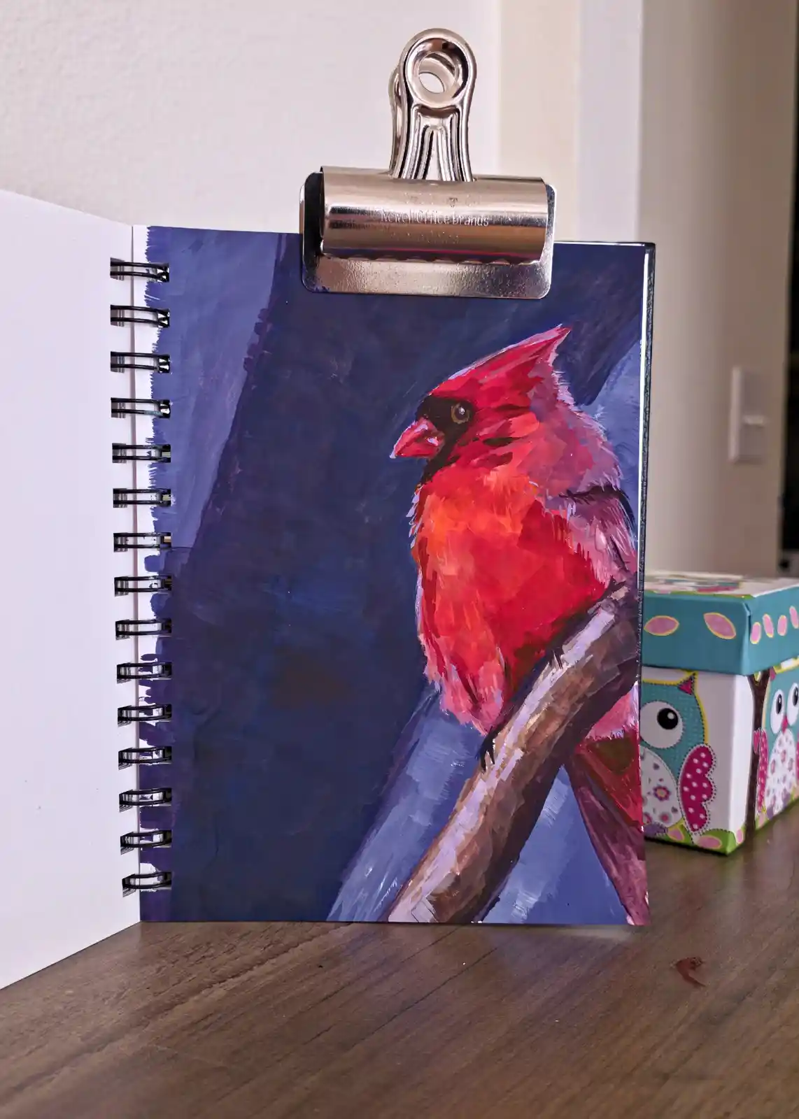 A spiral-bound notebook with a painted illustration of a red cardinal on its cover is clipped open with a silver metal clip…