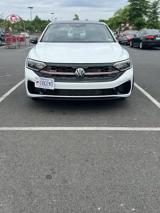 A white Volkswagen sedan is parked in a retail parking lot, facing the camera. Its Virginia license plate reads 'URGENT' in…