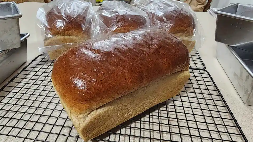A large, glossy loaf of bread sits prominently on a black wire cooling rack, with three smaller loaves wrapped in plastic…