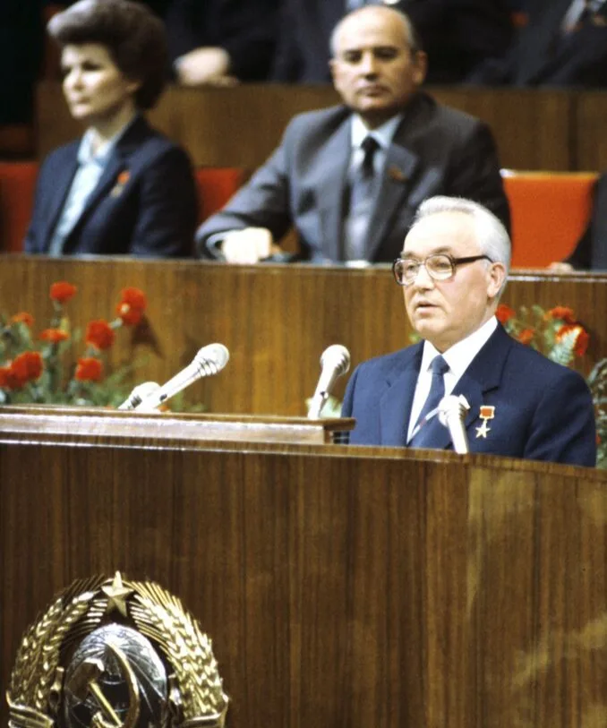 A man in a dark suit and glasses stands at a wooden podium adorned with a Soviet hammer and sickle emblem, speaking into…