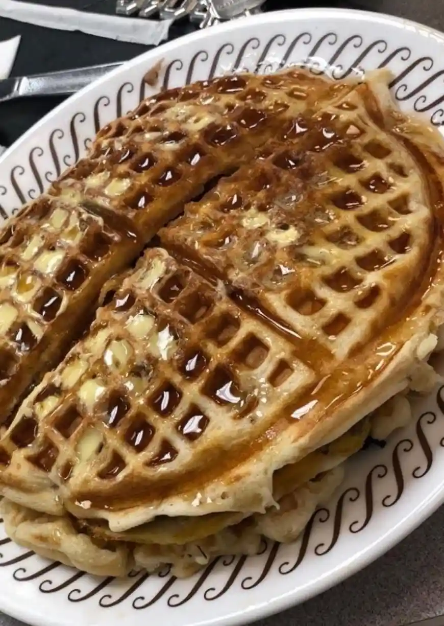 A close-up photo shows a tall stack of golden-brown waffles on a white plate with a brown swirling pattern, generously…