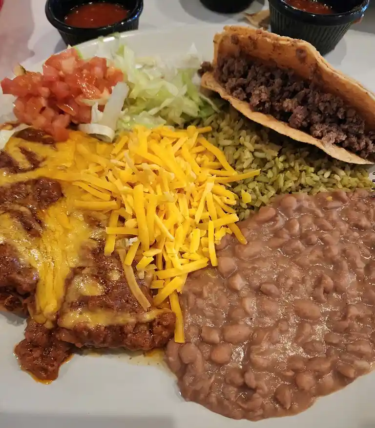 A close-up photo of a plate of Mexican food including refried beans, rice, shredded cheese, a taco, and toppings like…