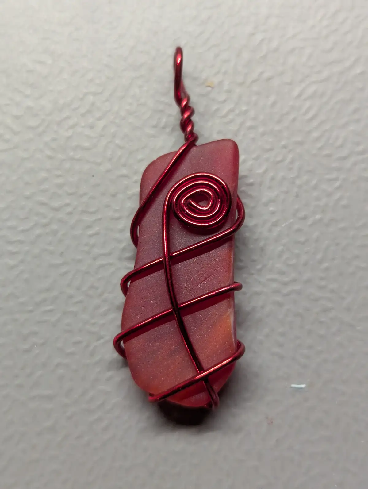 A handmade pendant featuring a translucent red glass piece wrapped in shiny red wire, with a spiral design at the top…