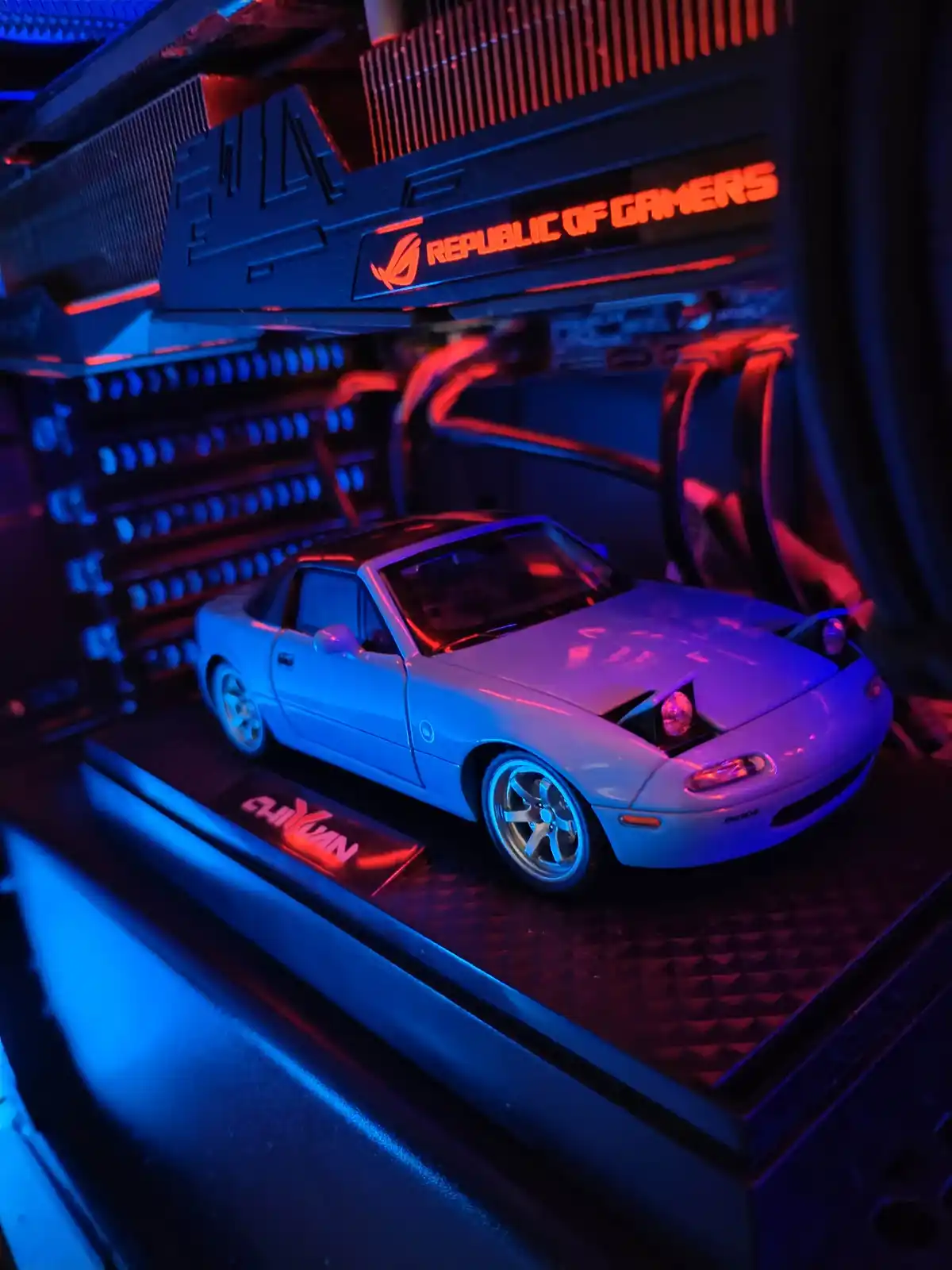 A small silver Mazda Miata model car sits inside a computer case, illuminated by blue and red LED lights. Behind it, a large…